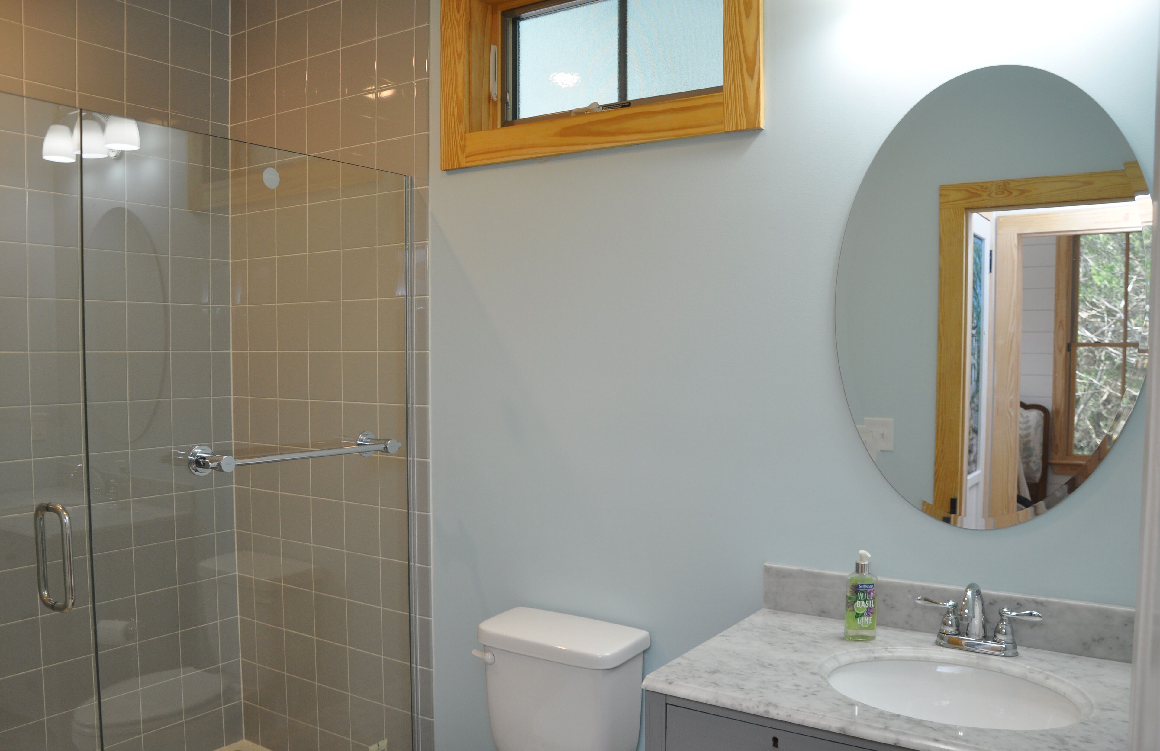 A bathroom with a glass shower enclosure, a toilet, a sink with a marble countertop, and a round mirror with a wooden frame.