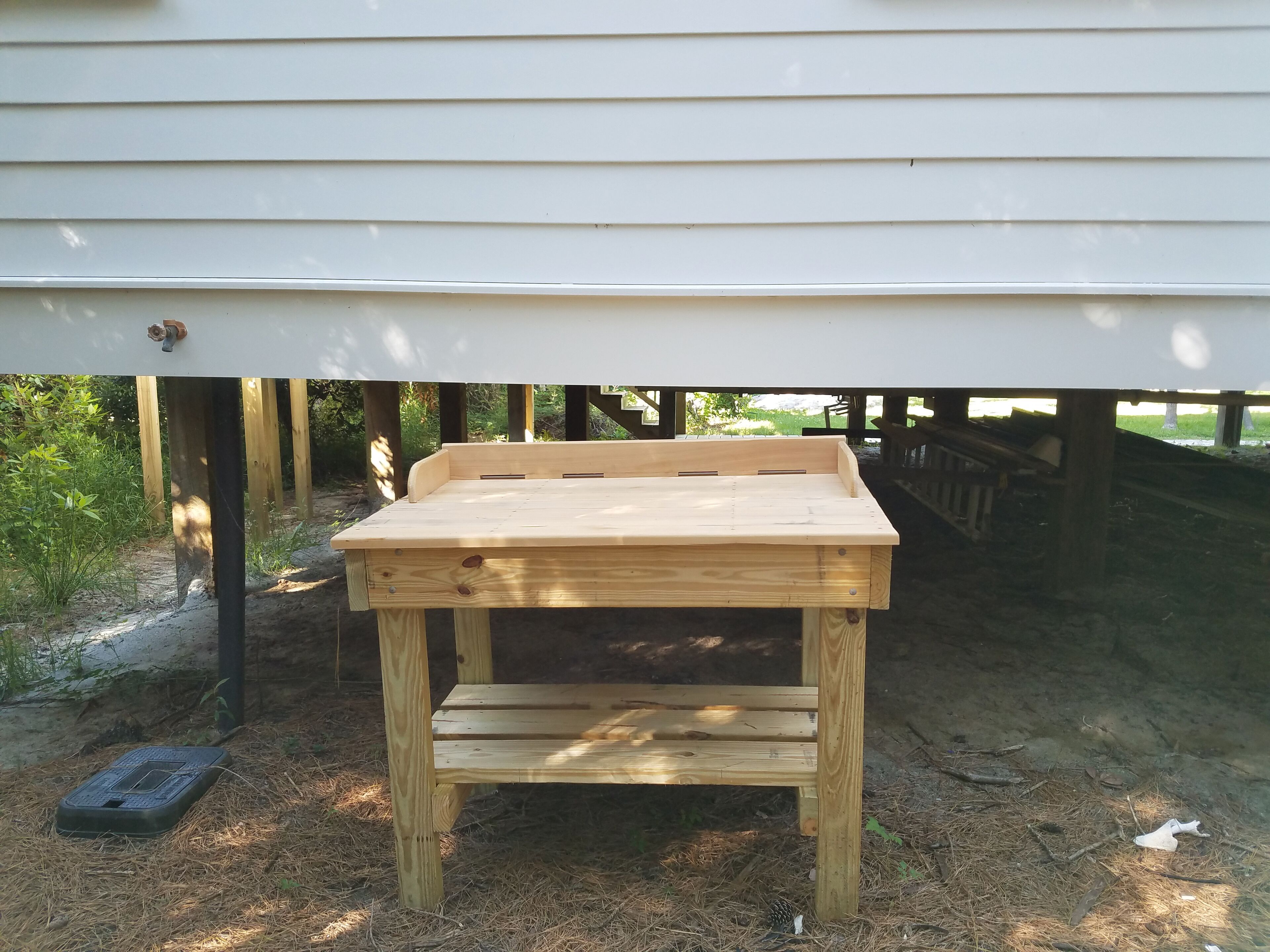 Een houten tafel onder een huis, met een metalen putdeksel in de buurt.
