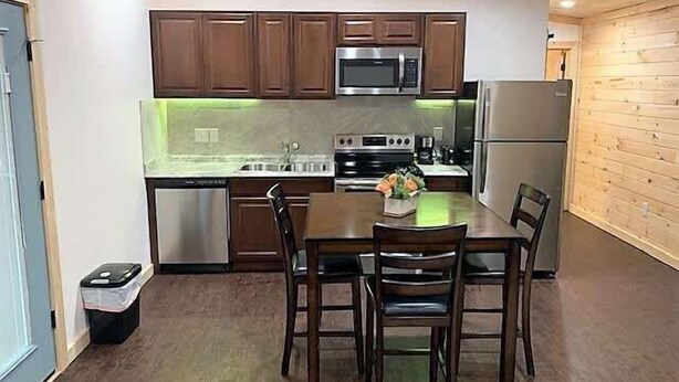 Une cuisine compacte avec des meubles en bois, un réfrigérateur en acier inoxydable, un four à micro-ondes et une cuisinière. On y trouve également une petite poubelle et une table avec des chaises.