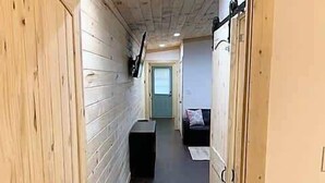 Un couloir étroit aux murs en bois et au plancher sombre, qui mène à une pièce contenant un lit et une fenêtre.