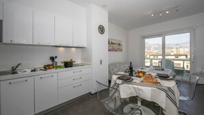 A modern kitchen with white cabinets, a dining area with a table set for two, and a large window offering a city view. - Sunrise House with Balcony and Parking, Lugano, Switzerland (Lugano)