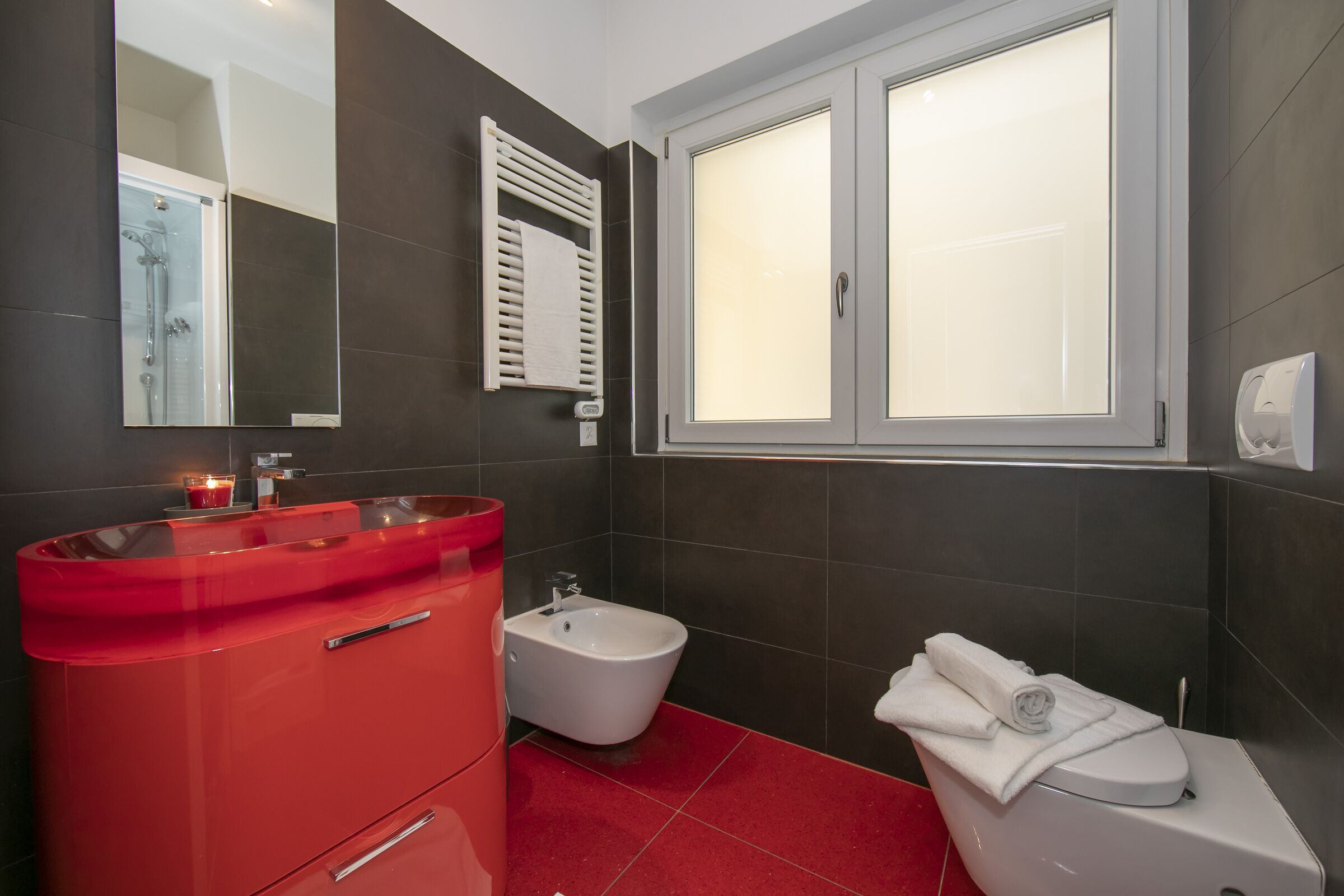 A modern bathroom with a red vanity, a bidet, a toilet with a towel, and a window with frosted glass.