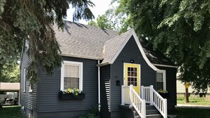 Une petite maison de plain-pied, à l’extérieur de couleur gris foncé, avec une porte d’entrée blanche et un petit porche doté d’une balustrade blanche.