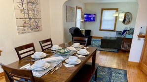 Un coin repas est aménagé avec une table en bois, dressée pour six personnes, et entourée de chaises. Une carte est accrochée au mur, et un téléviseur est fixé au-dessus d’un espace détente comprenant un canapé et des fauteuils.