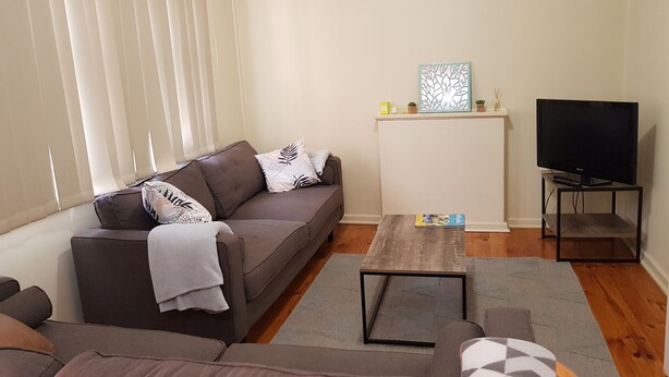 Una sala de estar con sofá, una mesita de centro, un televisor sobre un mueble bajo y un estante pequeño con adornos.