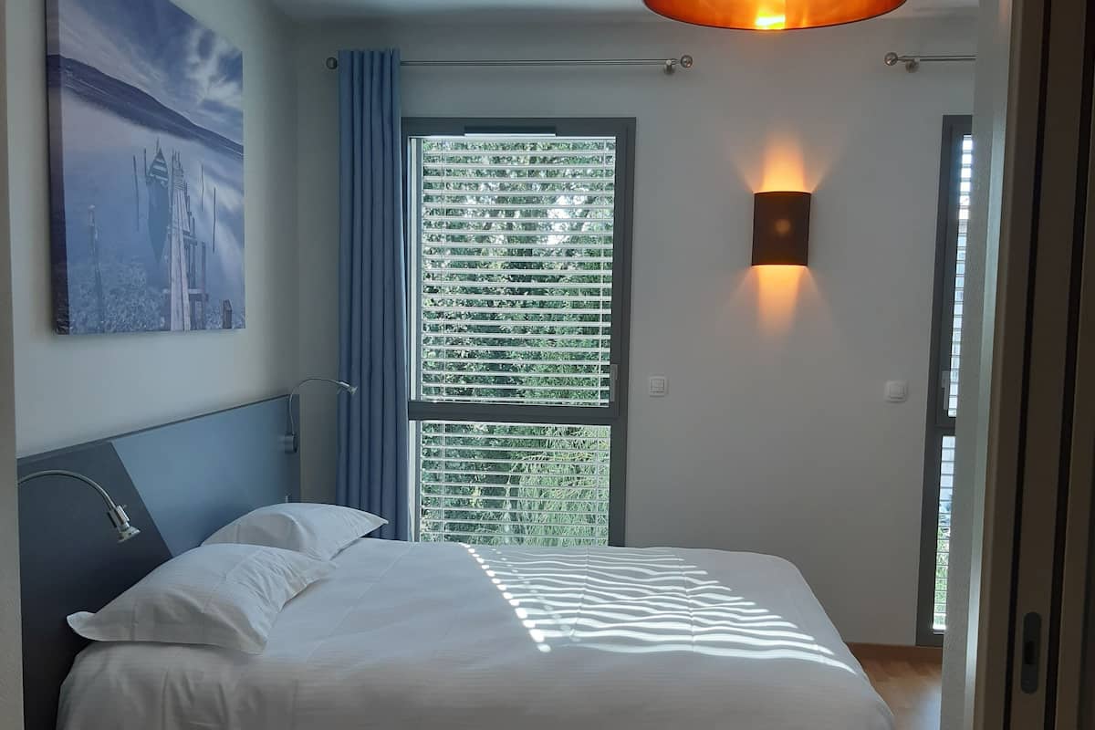 Ein modernes Schlafzimmer mit einem großen Bett, einer Kopfteil, einer Wandlampe, einem Fenster mit Jalousien und einem Gemälde an der Wand.