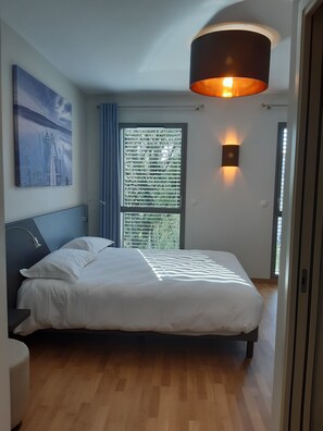 A modern bedroom with a large bed, a headboard, a wall-mounted lamp, a window with blinds, and a painting on the wall. - Hôtel du Parc (Lons-le-Saunier)