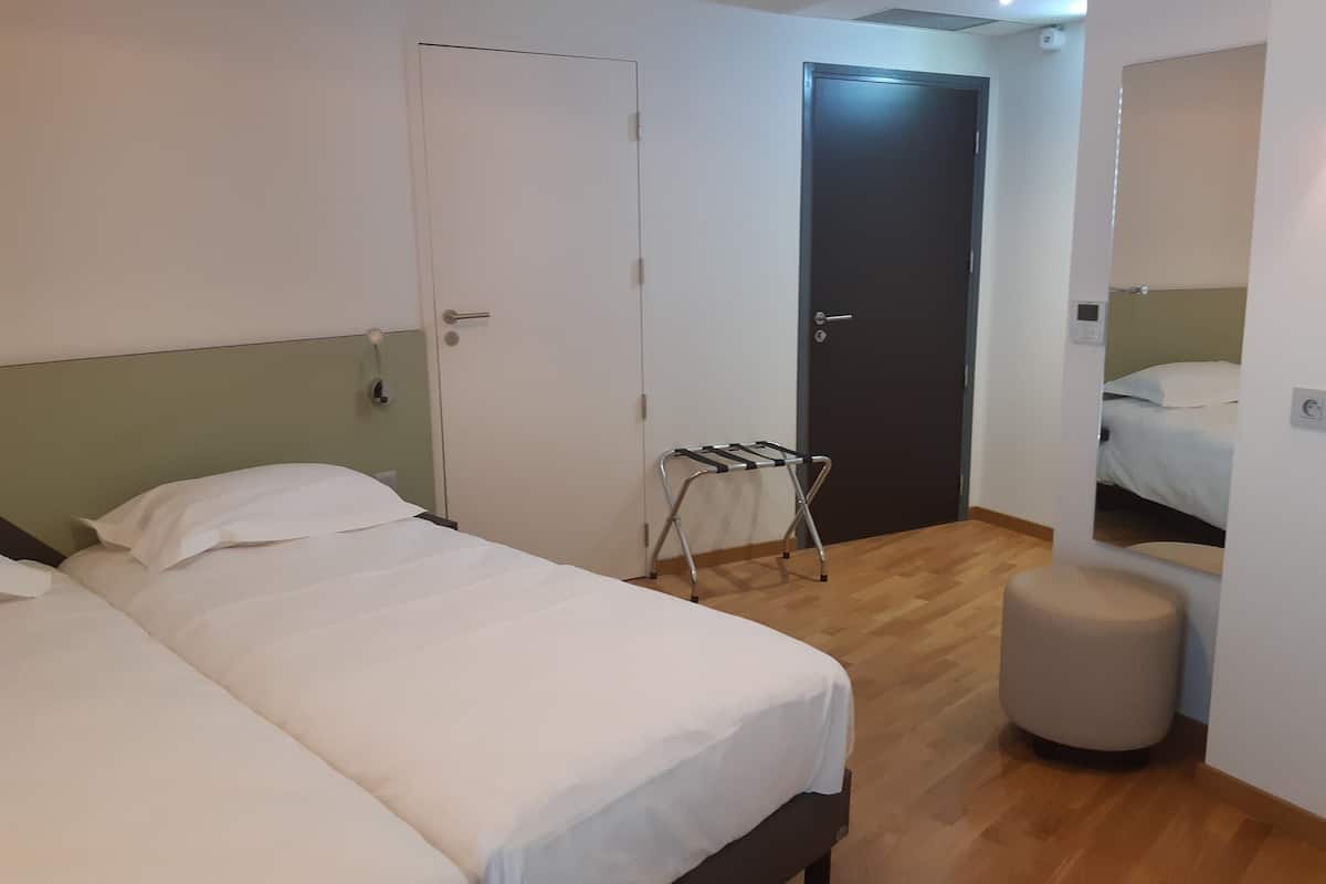 Ein Hotelzimmer mit zwei Betten, Holzboden, einem kleinen Hocker und einer Tür mit Handtuchhalter.