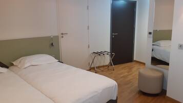 A hotel room with two beds, a wooden floor, a small stool, and a door with a towel rack.