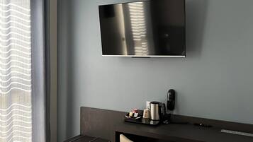 Une chambre moderne dotée d’un téléviseur à écran plat fixé au mur, d’un bureau sombre avec une chaise, et d’une petite table sur laquelle se trouvent une bouilloire et des tasses.