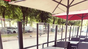 Een terras met tafels en rieten stoelen, grote ramen en een rode parasol.
