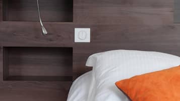 Ein Bett mit weißer Bettwäsche und einem orangefarbenen Kissen, eine weiße Lampe an einem hölzernen Kopfteil sowie eine Steckdose.
