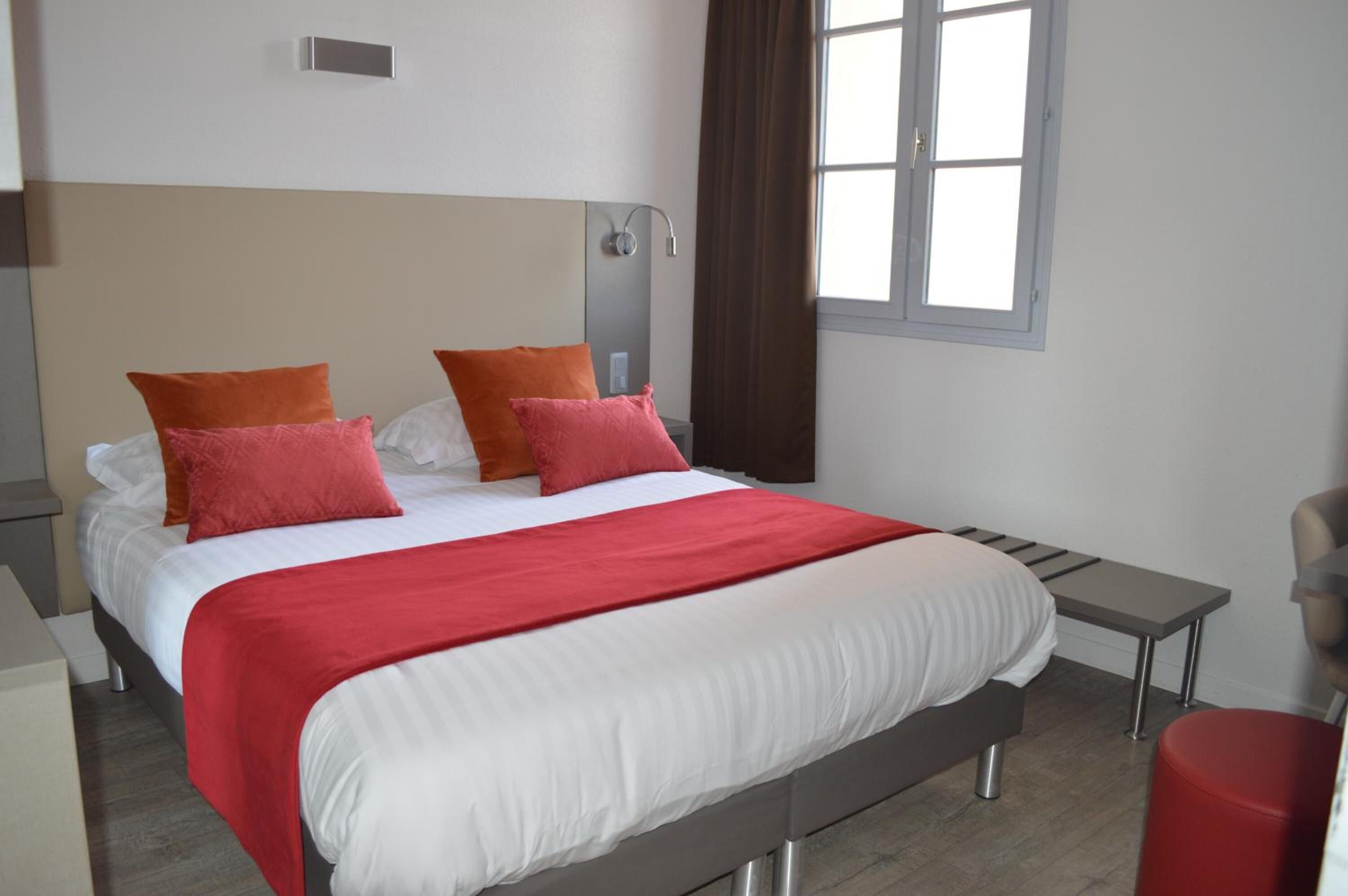 A modern hotel room with a large bed, two nightstands, a bedside table, a red bench, and a window with curtains.