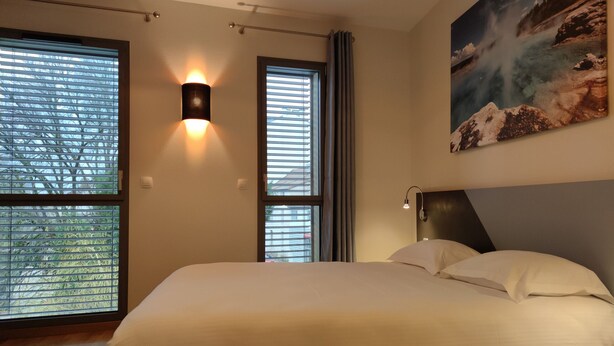 Een slaapkamer met een groot bed, twee ramen met jaloezieën, een wandlamp en een schilderij van een landschap.