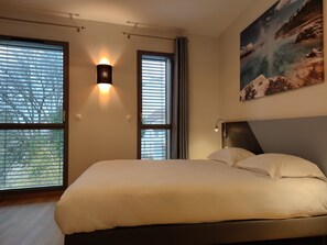 A bedroom with a large bed, two windows with blinds, a wall-mounted light, and a painting of a landscape. - Hôtel du Parc (Lons-le-Saunier)