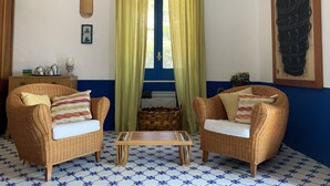 Una habitación con dos sillones de mimbre, una pequeña mesa de madera, un ventanal con cortinas verdes, un aire acondicionado instalado en la pared y una obra de arte enmarcada.