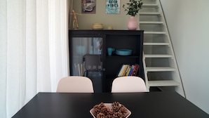 Un coin repas avec une table noire, deux chaises blanches, une assiette remplie de pommes de pin, une bibliothèque avec des livres et des objets décoratifs, un vase rose et un escalier.