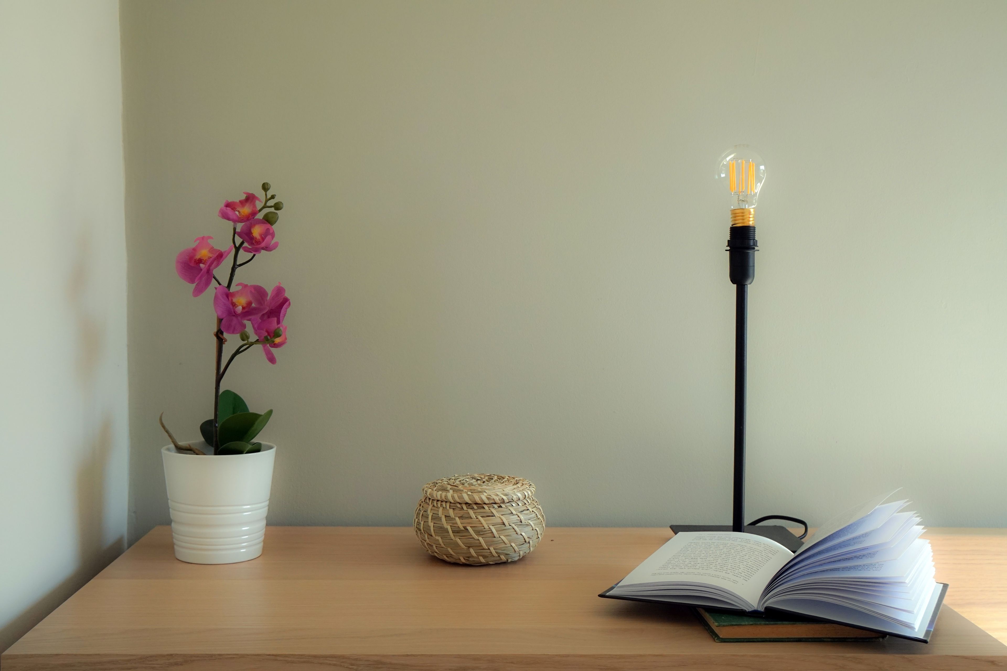 Una mesa de madera con una orquídea blanca en maceta, una cesta tejida y un libro abierto bajo una lámpara de pie.