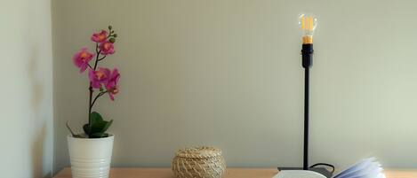 Una mesa de madera con una orquídea blanca en maceta, una cesta tejida y un libro abierto bajo una lámpara de pie.