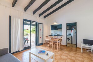 Una sala de estar con sofá, una mesa con un libro y control remoto, un comedor con sillas, una cocina con electrodomésticos y un ventanal corredizo que da al exterior.