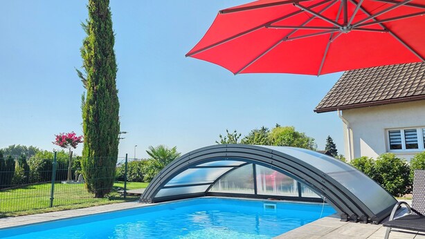 A swimming pool with a modern design, surrounded by a fence, a tall tree, and a red umbrella. - chez moumie (La Grande-Béroche)