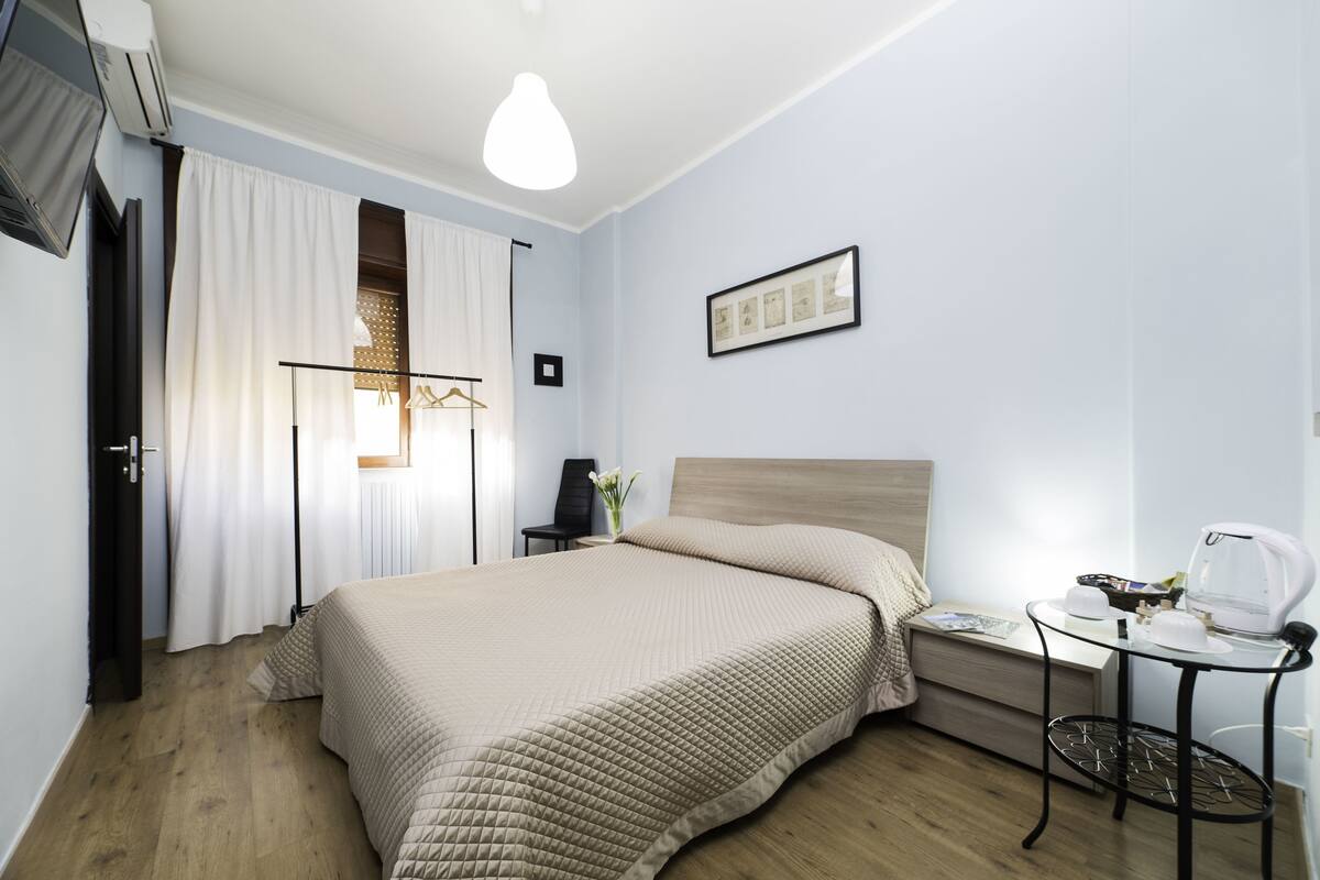 A neatly arranged hotel room with a bed, bedside table, and a small table with a kettle.