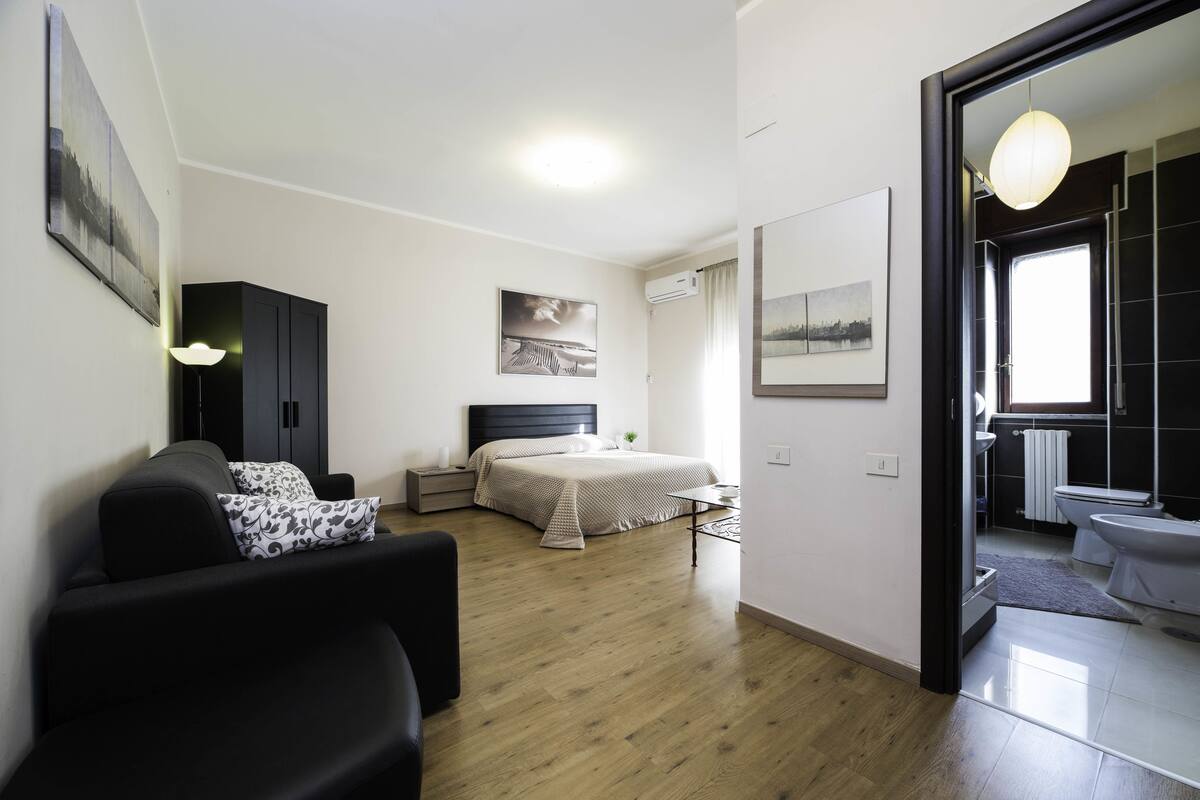 A modern bedroom with a bed, bedside table, and a nightstand. There is a sofa in the foreground and a bathroom visible through an open door.