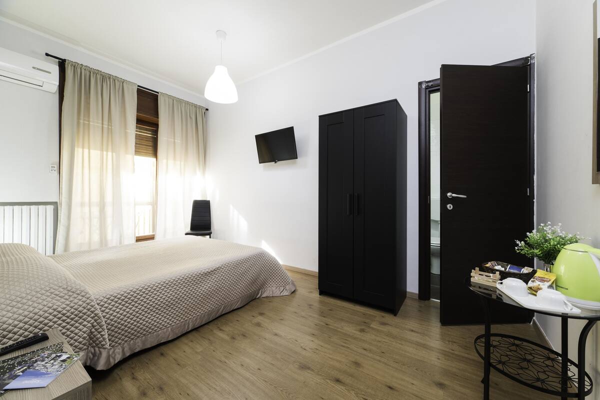 A bedroom with a bed, a wardrobe, a television, and a table with a lamp and some items on it.