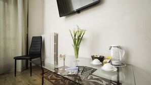 Une pièce moderne avec une table en verre, une chaise noire, un radiateur blanc, un téléviseur fixé au mur, un vase avec des fleurs blanches, une machine à café et un petit objet décoratif.