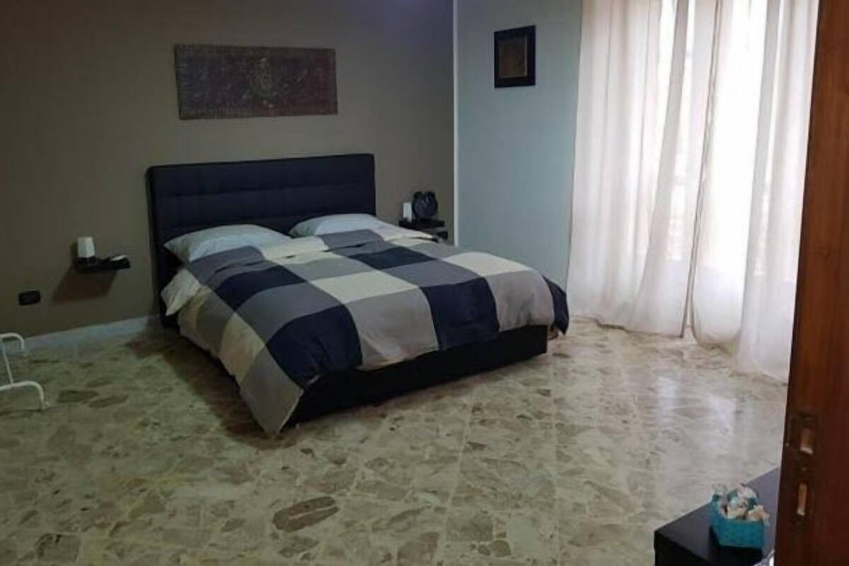 Un dormitorio con cama, mesita de noche, un reloj en la pared y una ventana con cortinas.