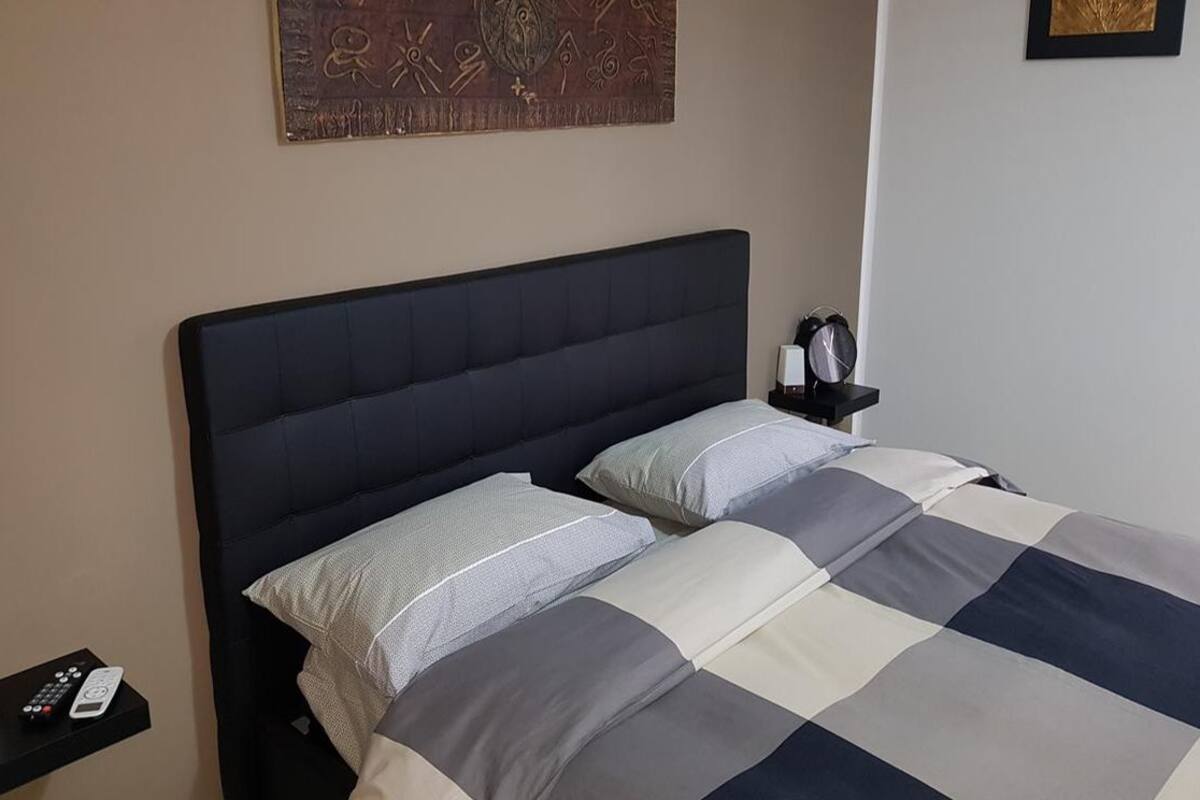 Una cama bien hecha con colcha a cuadros y almohadas, una mesita de noche con un control remoto, y una obra de arte en la pared sobre la cama.