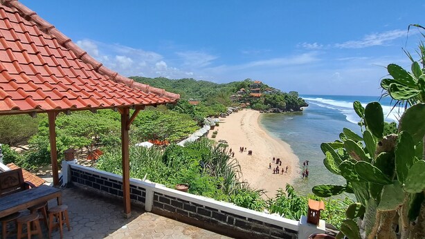 A beach with a red-tiled roof structure, a sandy shore, and lush greenery. - Omah Sundak Homestay (Wonosari)