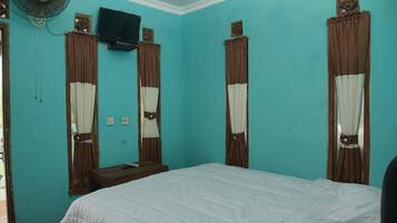 Une chambre avec des murs de couleur turquoise, un lit recouvert d’une literie blanche, un téléviseur fixé au-dessus d’une table de chevet en bois, et des rideaux à panneaux marron et blancs.