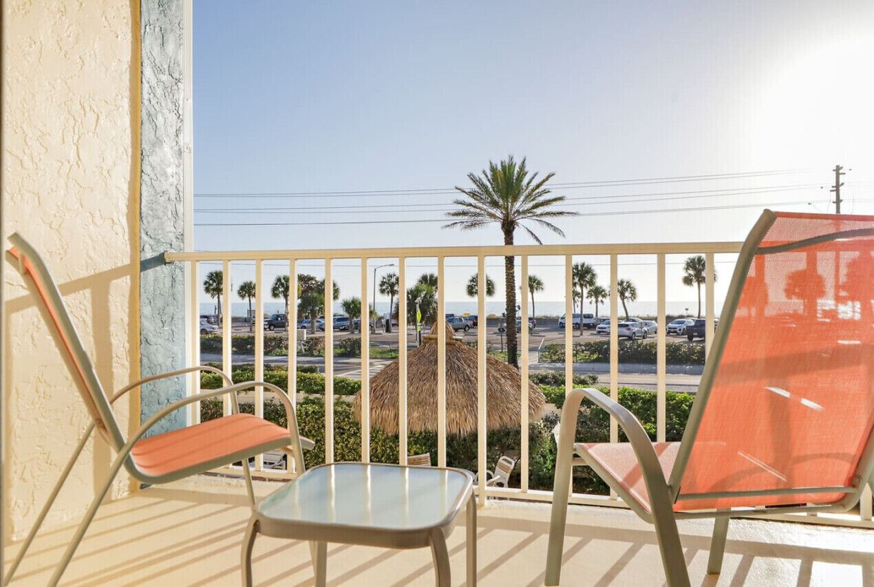 A balcony with two chairs and a table, overlooking a street with palm trees and parked cars.