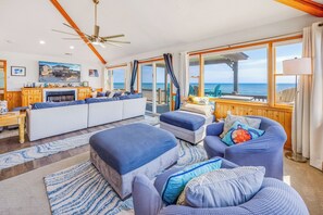 En rummelig opholdsstue med en stor sofa, en mindre sofa og et spisebord. Store vinduer giver udsigt til havet.