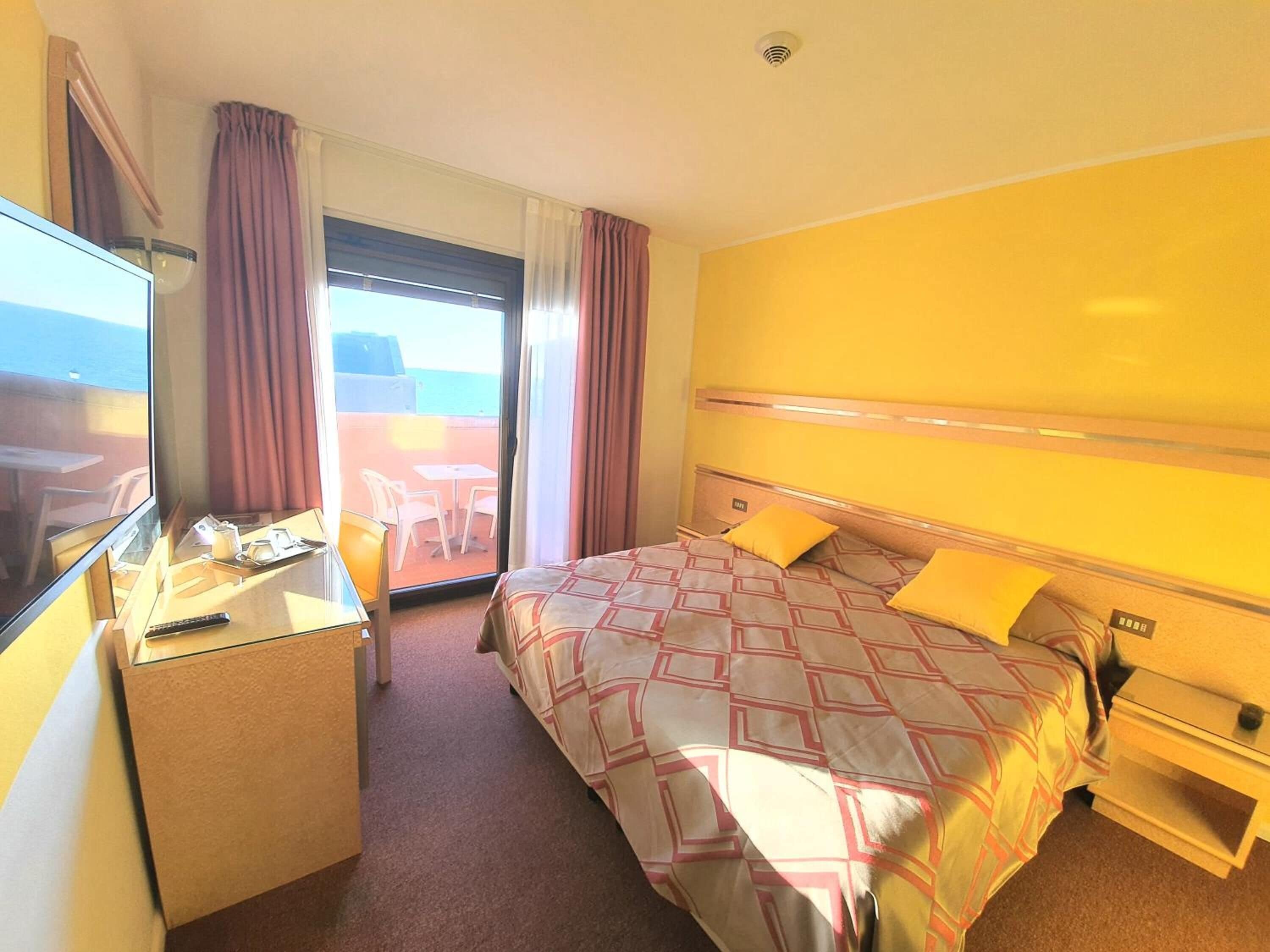 A hotel room with a bed, a desk, and a chair. There is a window with curtains and a view of the outside.