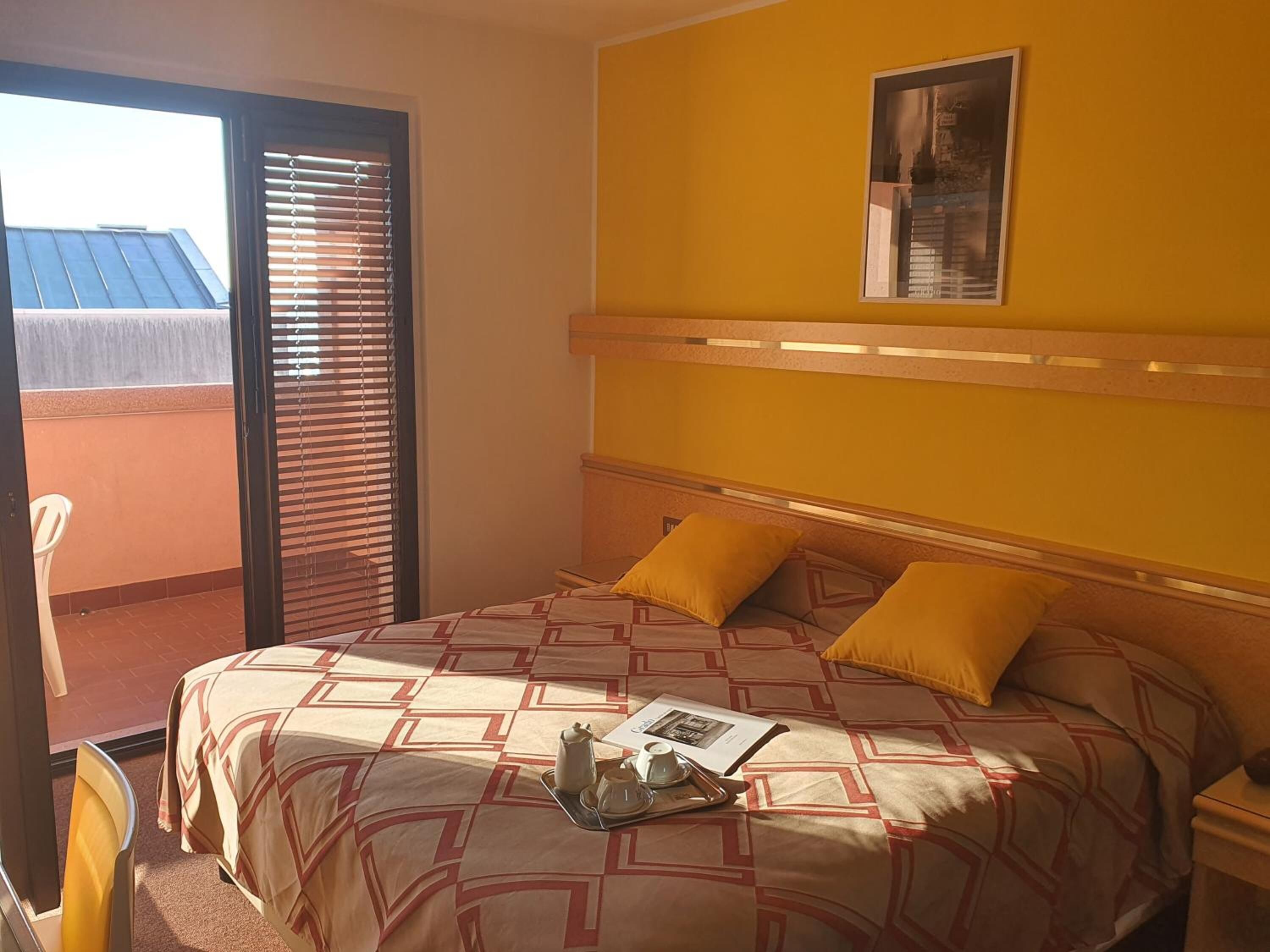 A hotel room with a bed, two pillows, a nightstand, a window with blinds, and a view of a building outside.
