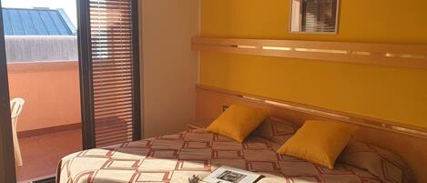 A hotel room with a bed, two pillows, a nightstand, a window with blinds, and a view of a building outside.