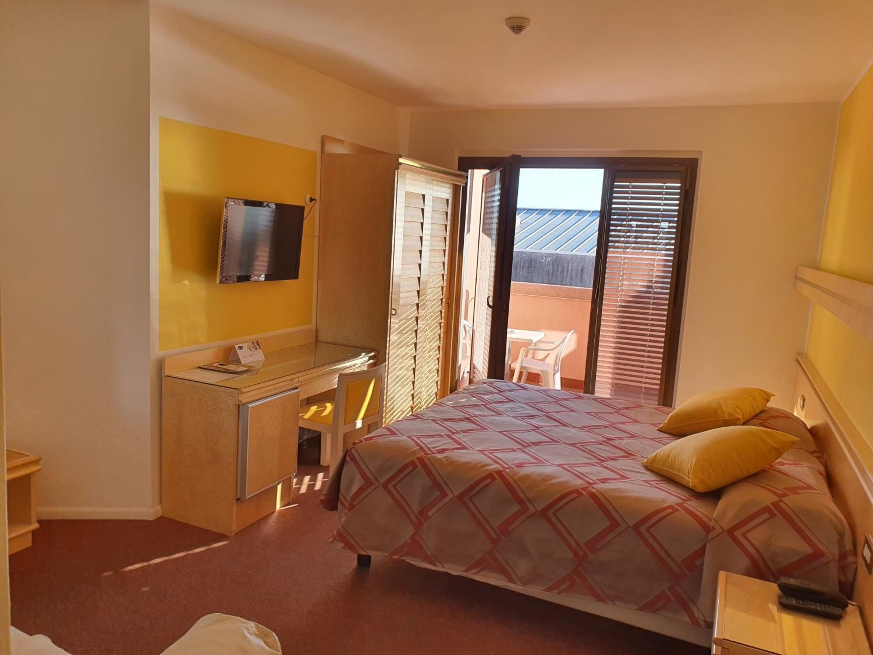 Habitación de hotel con cama, escritorio con televisor, una mesita pequeña y puerta corrediza a un espacio exterior.