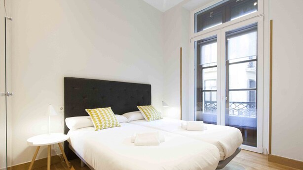 A modern hotel room with a large bed, two bedside tables, and a view of the outside balcony through a glass door. - Dream | The Rentals Collection (San Sebastián/Donostia)
