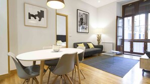 A modern dining area with a round table, grey chairs, and a white tablecloth. There is a grey sofa with yellow cushions in the background, a large window with a view of buildings, and a floor lamp. - Dream | The Rentals Collection (San Sebastián/Donostia)
