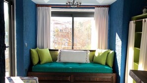 Une pièce comprenant un banc intégré avec des coussins, une chaise en bois, un lit recouvert d’une couverture colorée et une fenêtre avec des rideaux.
