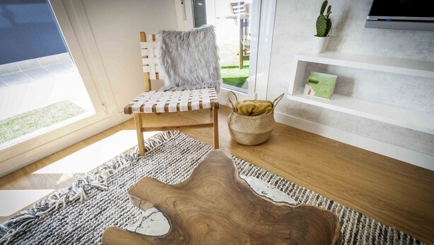 A modern living room with a wooden coffee table, a cushioned chair, a woven basket, and a shelf with a cactus and a book. - Bellagio | The Rentals Collection (San Sebastián)