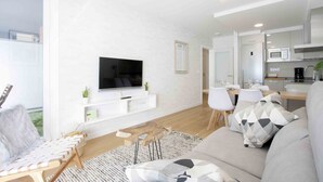 A modern living room with a flat-screen TV, a sofa, a coffee table, and a dining area with chairs and a table. - Bellagio | The Rentals Collection (San Sebastián)