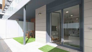 A modern building with a glass door entrance, a small outdoor seating area with a table and chairs, and a grassy patch. - Bellagio | The Rentals Collection (San Sebastián)