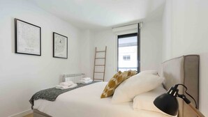 A neatly made bed with white linens and a gray throw, a black desk lamp, and two framed maps on the wall. - Bellagio | The Rentals Collection (San Sebastián)