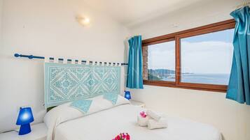 Ein Schlafzimmer mit einem Bett, einem Nachttisch, einer Lampe, einem Fenster mit Vorhängen und Blick aufs Meer.
