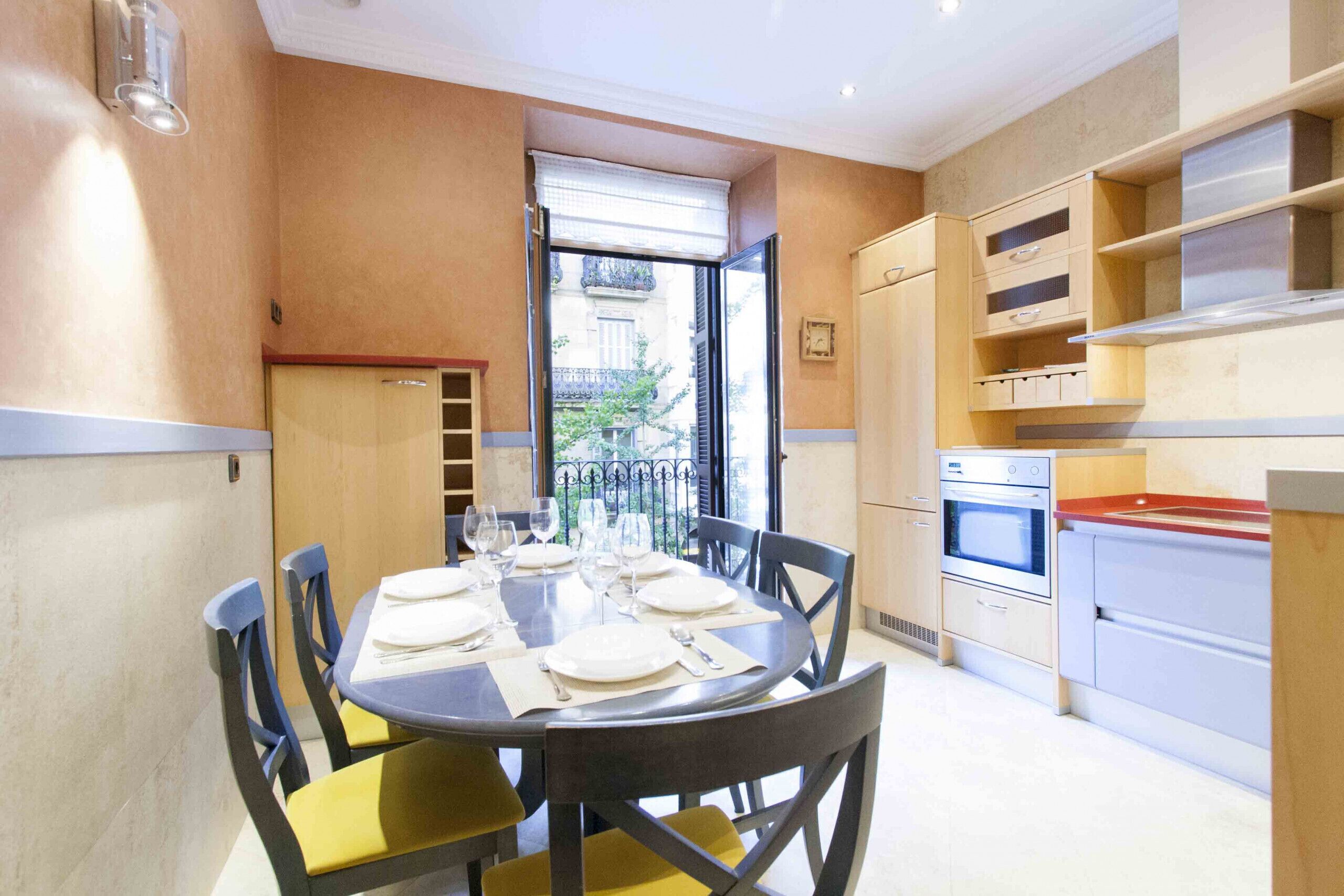 A dining area with a round table set for four, a kitchen with modern appliances, and a view through an open door.