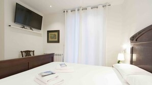 A hotel room with a bed, a flat-screen TV mounted on the wall, a framed picture, a window with white curtains, and a bedside lamp. - Gallery | The Rentals Collection (San Sebastián/Donostia)