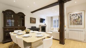 A dining area with a wooden table set for six, white chairs, a dark wooden cabinet with glass doors, a fireplace, and a painting on the wall. - Gallery | The Rentals Collection (San Sebastián/Donostia)